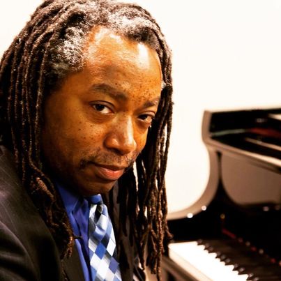 Wendel Werner sits at the piano, his hair is in thin dreadlocks, and he is wearing a black suit with a checked blue tie